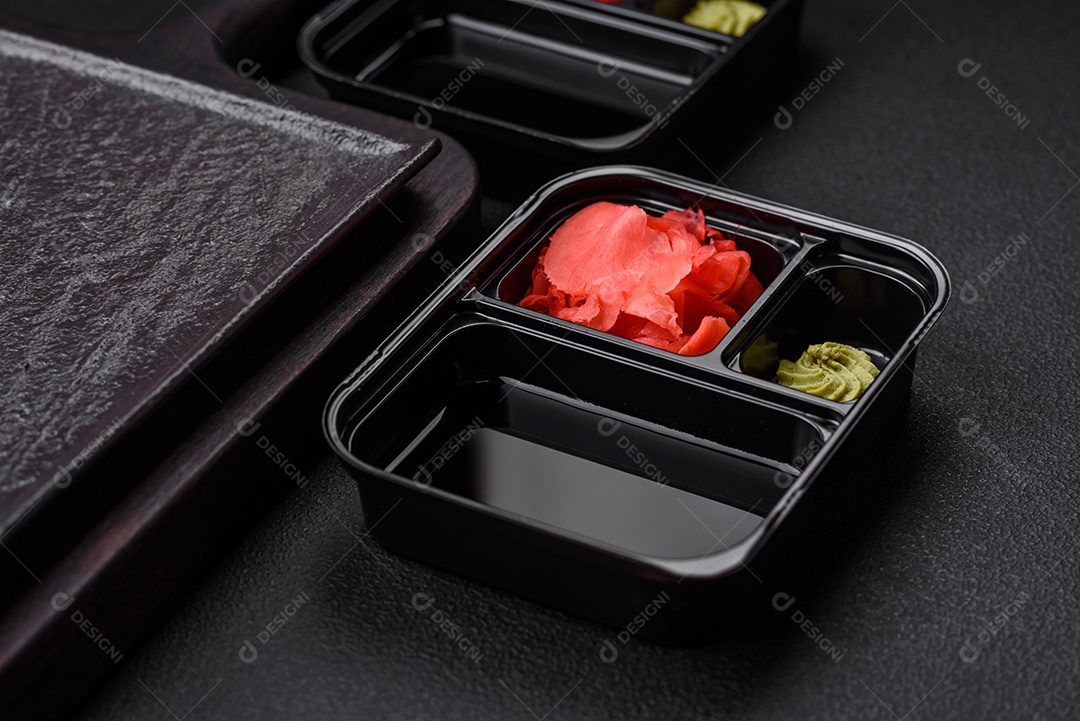 Gengibre em conserva, molho de soja e wasabi em recipientes de plástico porcionados sobre um fundo escuro de concreto