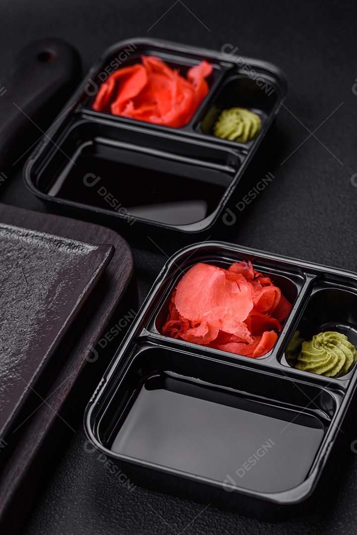 Gengibre em conserva, molho de soja e wasabi em recipientes de plástico porcionados sobre um fundo escuro de concreto
