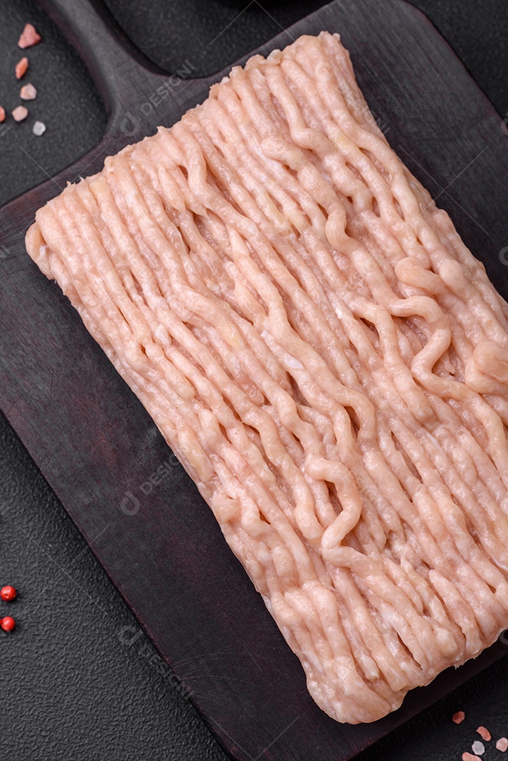 Frango picado fresco com sal, especiarias e ervas em uma tábua de madeira sobre um fundo escuro de concreto