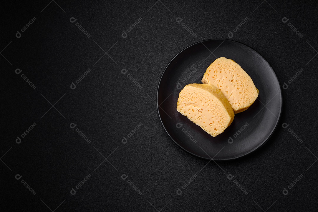 Delicioso queijo amarelo poroso cortado em pedaços grandes numa placa cerâmica sobre um fundo escuro de concreto