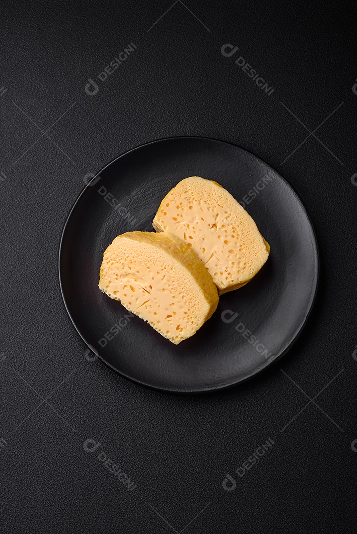 Delicioso queijo amarelo poroso cortado em pedaços grandes numa placa cerâmica sobre um fundo escuro de concreto