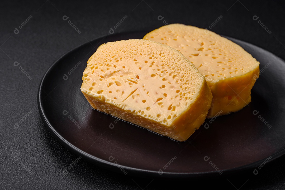 Delicioso queijo amarelo poroso cortado em pedaços grandes numa placa cerâmica sobre um fundo escuro de concreto