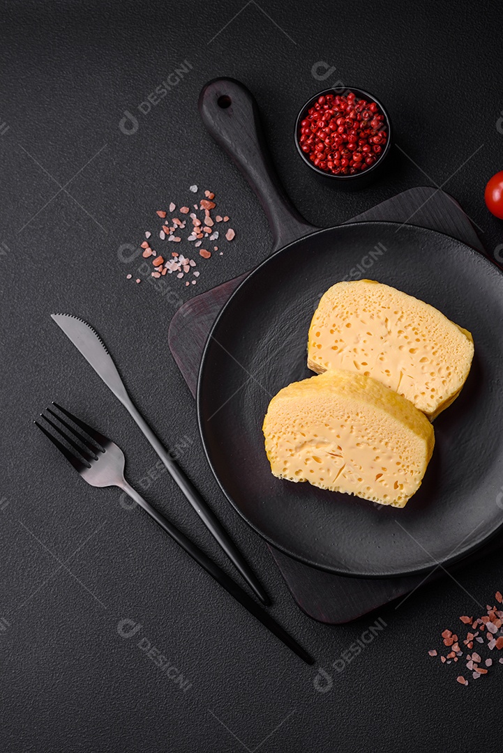 Delicioso queijo amarelo poroso cortado em pedaços grandes numa placa cerâmica sobre um fundo escuro de concreto