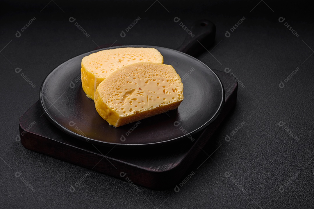 Delicioso queijo amarelo poroso cortado em pedaços grandes numa placa cerâmica sobre um fundo escuro de concreto