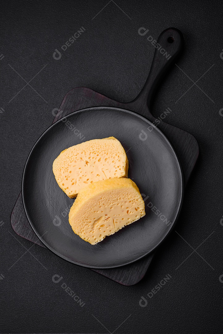 Delicioso queijo amarelo poroso cortado em pedaços grandes numa placa cerâmica sobre um fundo escuro de concreto