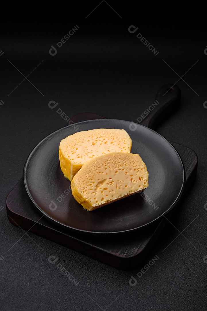 Delicioso queijo amarelo poroso cortado em pedaços grandes numa placa cerâmica sobre um fundo escuro de concreto