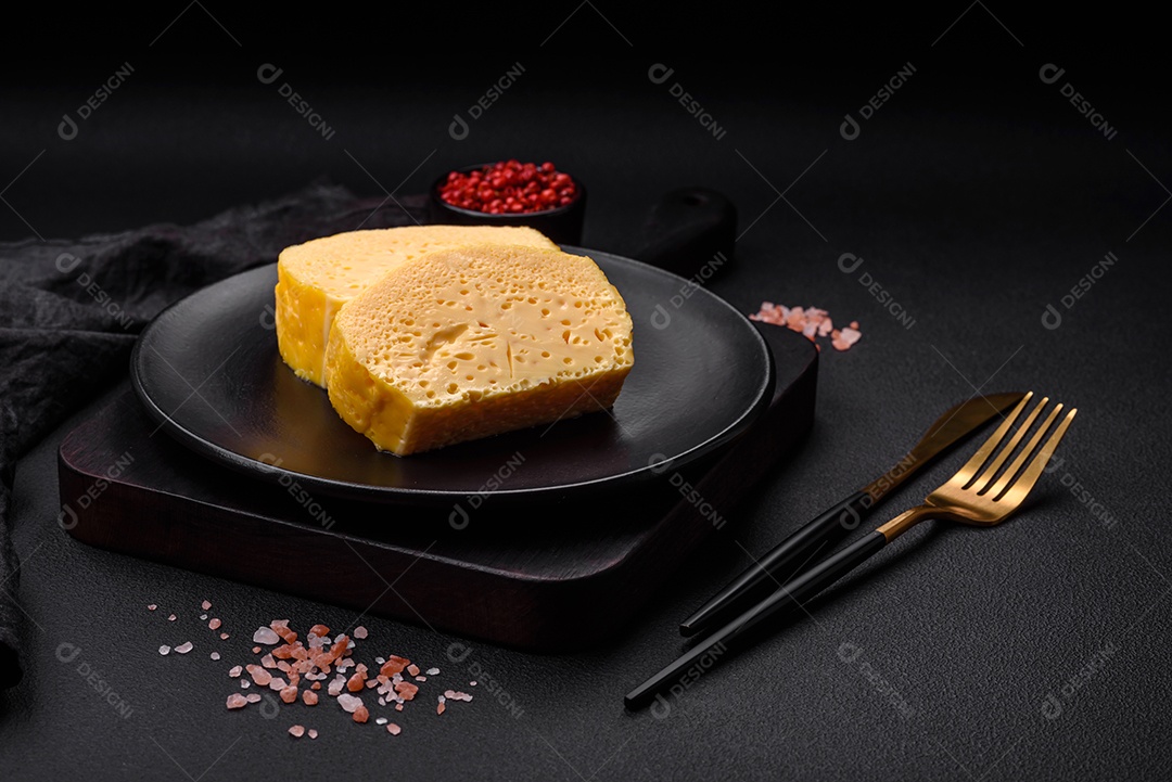 Delicioso queijo amarelo poroso cortado em pedaços grandes numa placa cerâmica sobre um fundo escuro de concreto