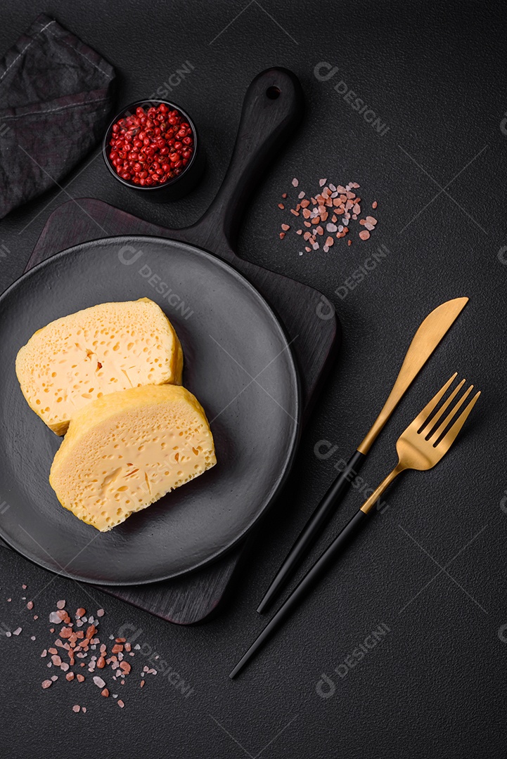 Delicioso queijo amarelo poroso cortado em pedaços grandes numa placa cerâmica sobre um fundo escuro de concreto