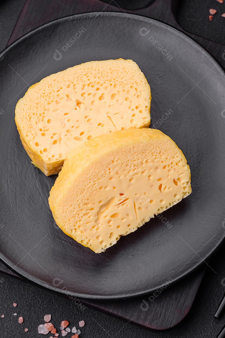 Delicioso queijo amarelo poroso cortado em pedaços grandes numa placa cerâmica sobre um fundo escuro de concreto