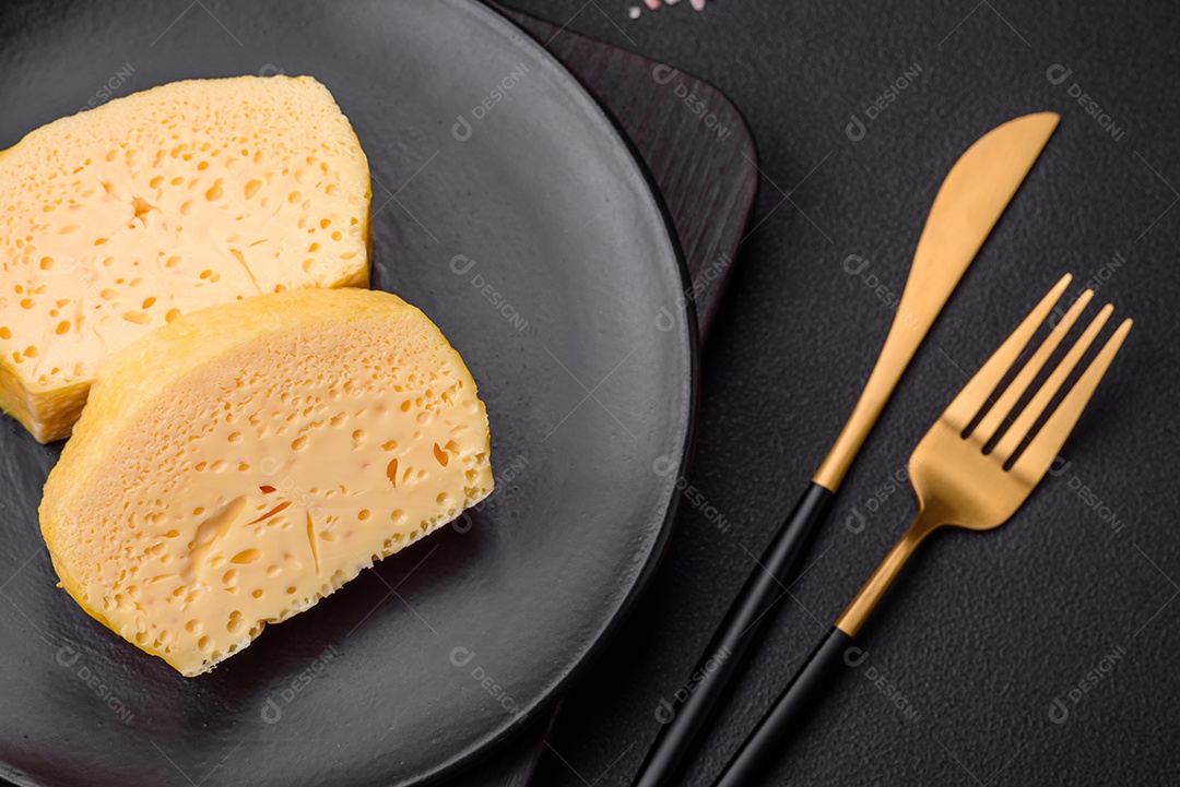 Delicioso queijo amarelo poroso cortado em pedaços grandes numa placa cerâmica sobre um fundo escuro de concreto