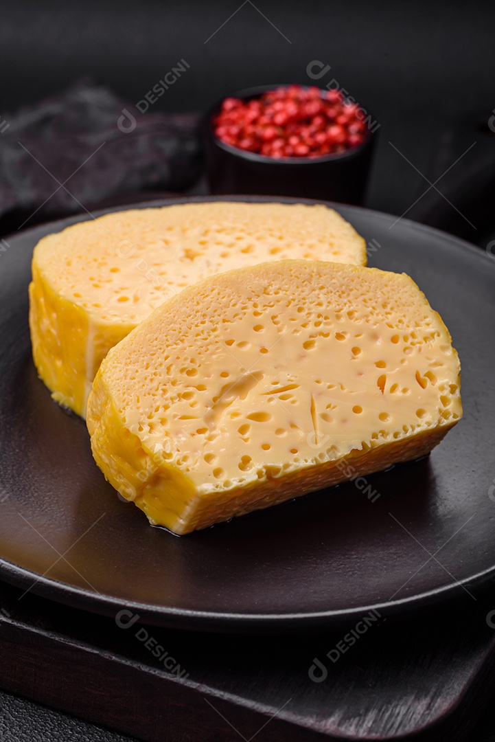 Delicioso queijo amarelo poroso cortado em pedaços grandes numa placa cerâmica sobre um fundo escuro de concreto