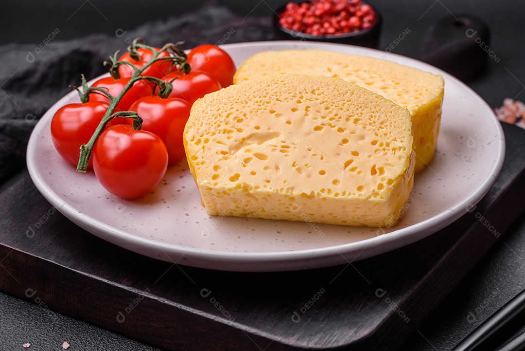 Delicioso queijo amarelo poroso cortado em pedaços grandes numa placa cerâmica sobre um fundo escuro de concreto