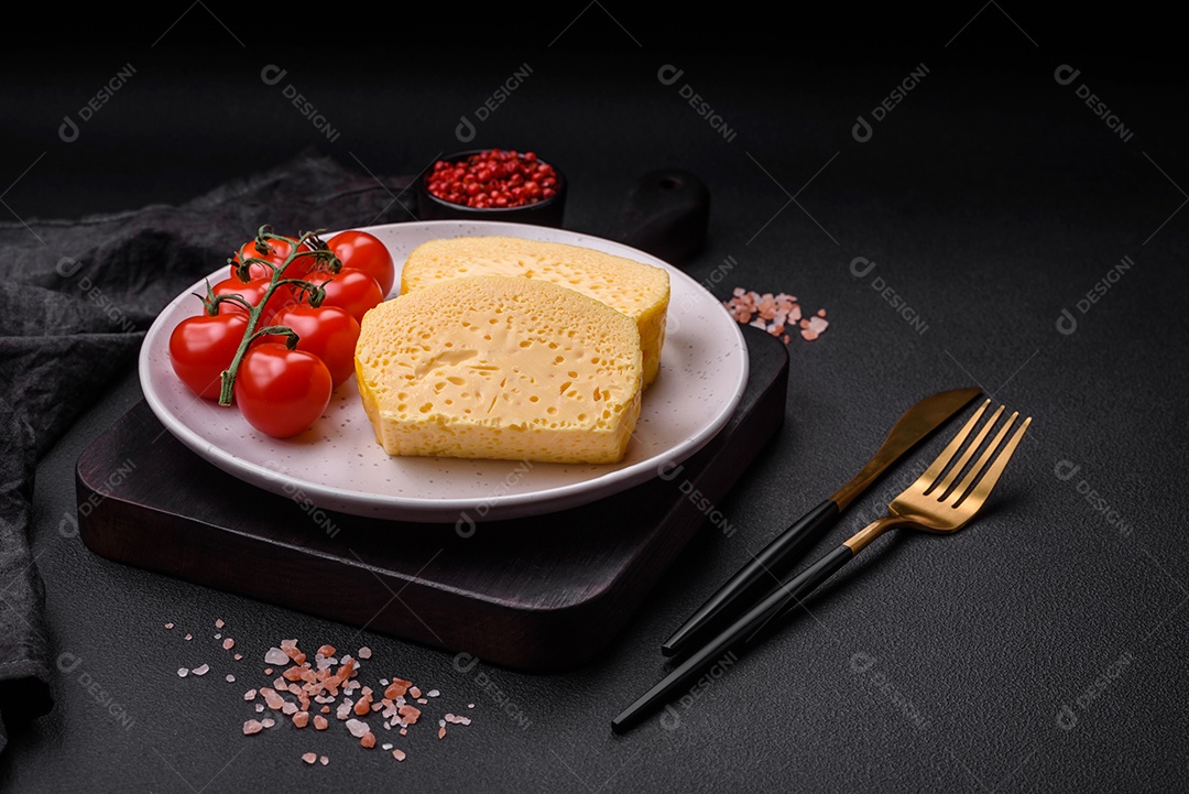 Delicioso queijo amarelo poroso cortado em pedaços grandes numa placa cerâmica sobre um fundo escuro de concreto