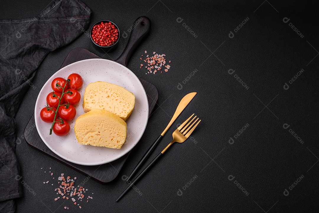 Delicioso queijo amarelo poroso cortado em pedaços grandes numa placa cerâmica sobre um fundo escuro de concreto