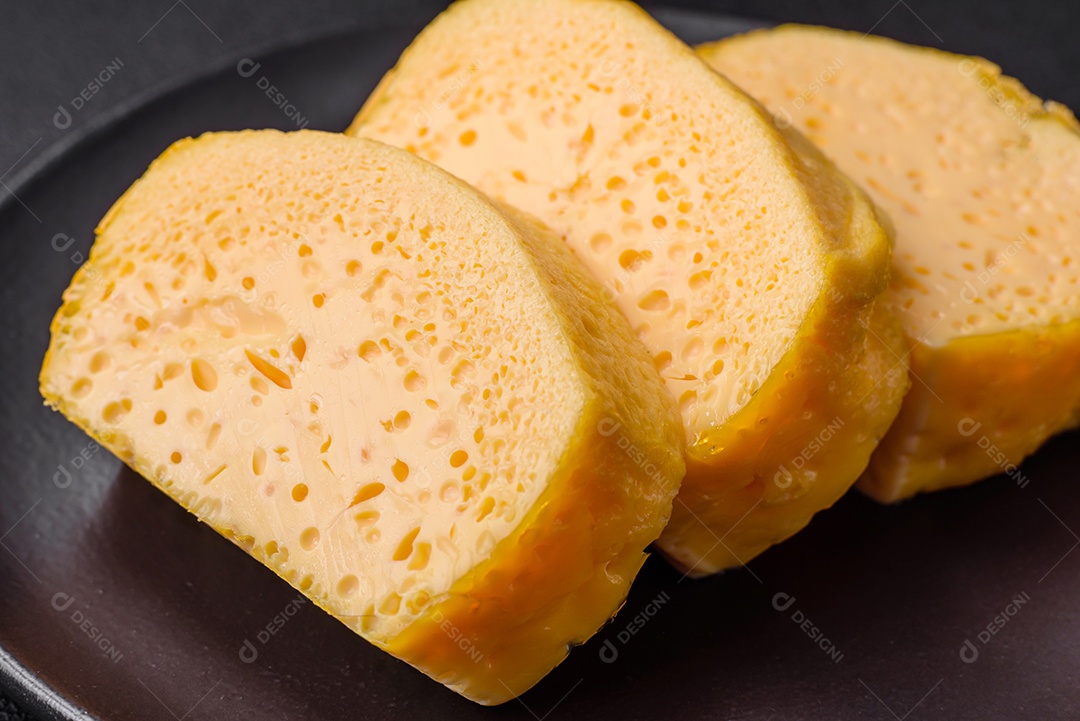 Delicioso queijo amarelo poroso cortado em pedaços grandes numa placa cerâmica sobre um fundo escuro de concreto