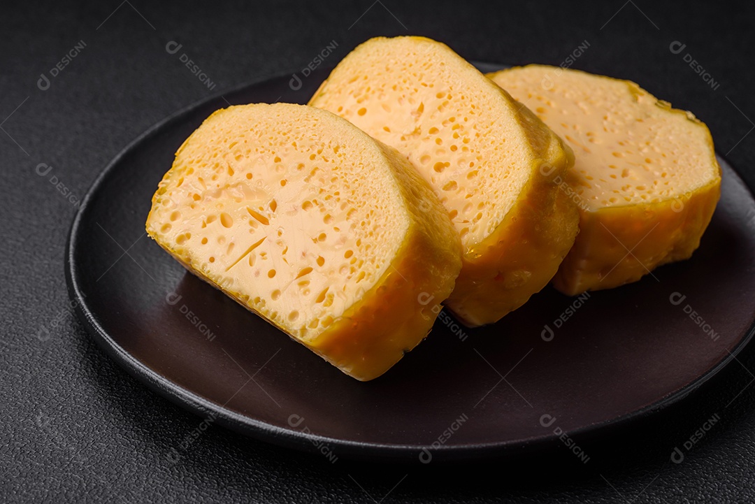 Delicioso queijo amarelo poroso cortado em pedaços grandes numa placa cerâmica sobre um fundo escuro de concreto