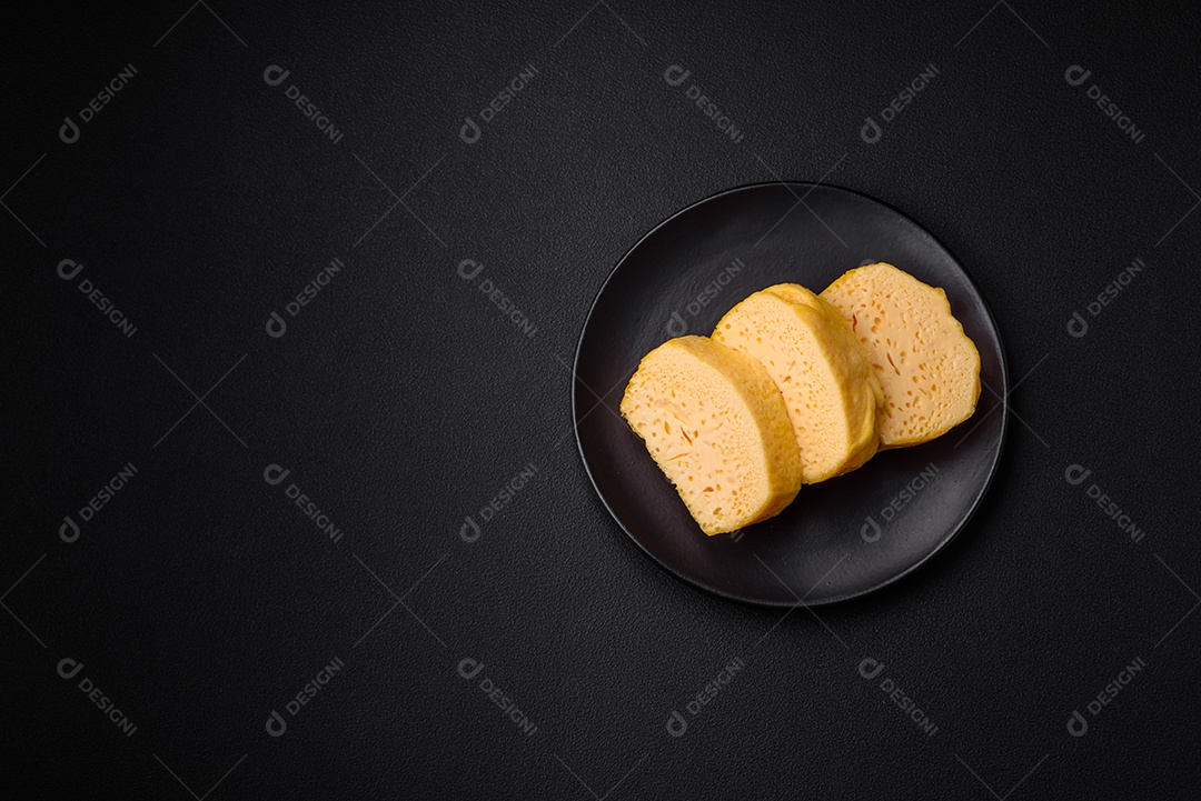 Delicioso queijo amarelo poroso cortado em pedaços grandes numa placa cerâmica sobre um fundo escuro de concreto