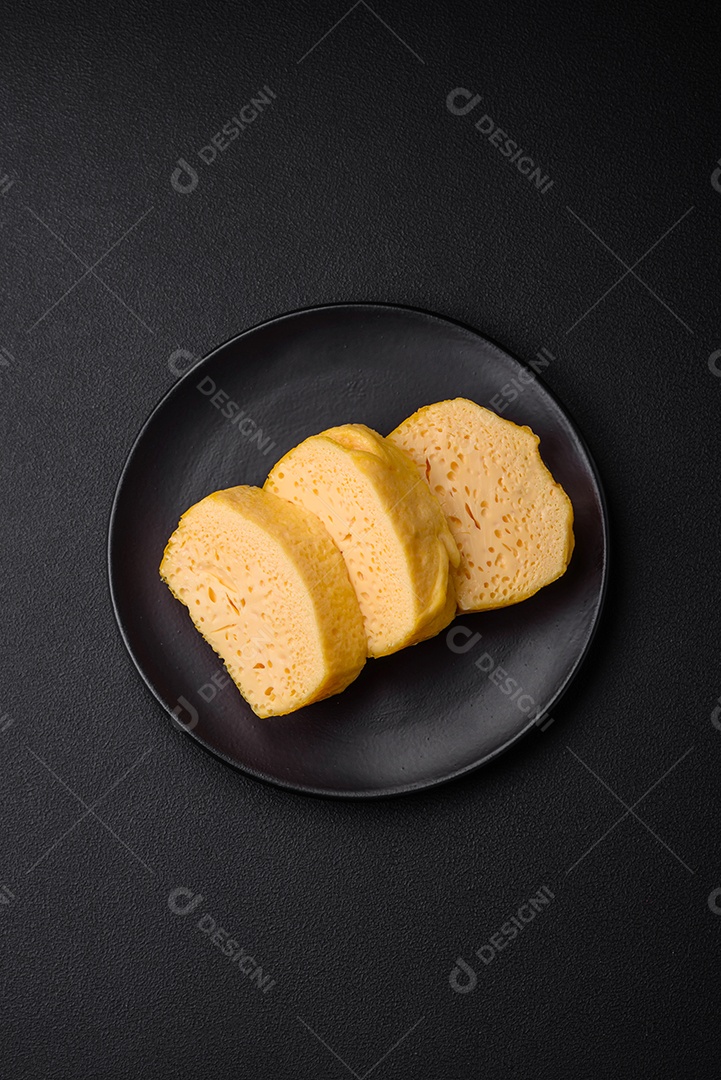 Delicioso queijo amarelo poroso cortado em pedaços grandes numa placa cerâmica sobre um fundo escuro de concreto
