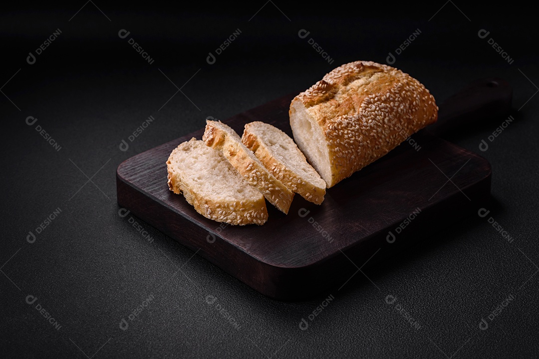 Baguete de farinha de trigo crocante com sementes de gergelim em uma tábua de madeira sobre um fundo escuro de concreto