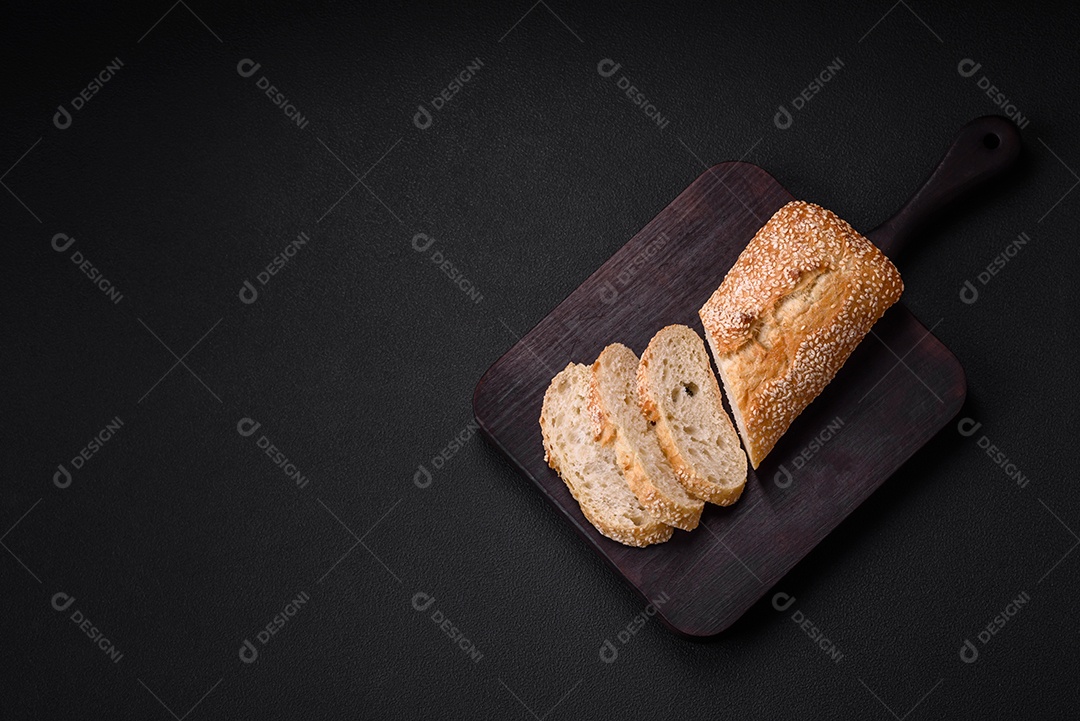 Baguete de farinha de trigo crocante com sementes de gergelim em uma tábua de madeira sobre um fundo escuro de concreto