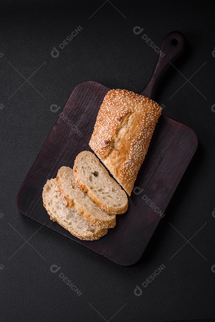 Baguete de farinha de trigo crocante com sementes de gergelim em uma tábua de madeira sobre um fundo escuro de concreto