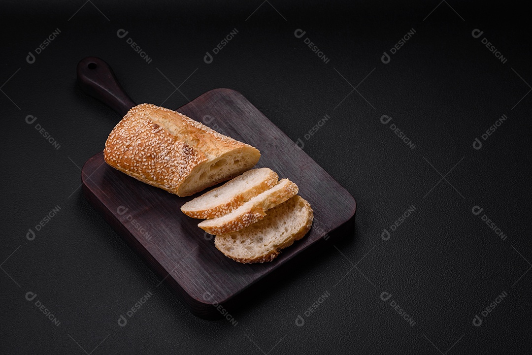 Baguete de farinha de trigo crocante com sementes de gergelim em uma tábua de madeira sobre um fundo escuro de concreto