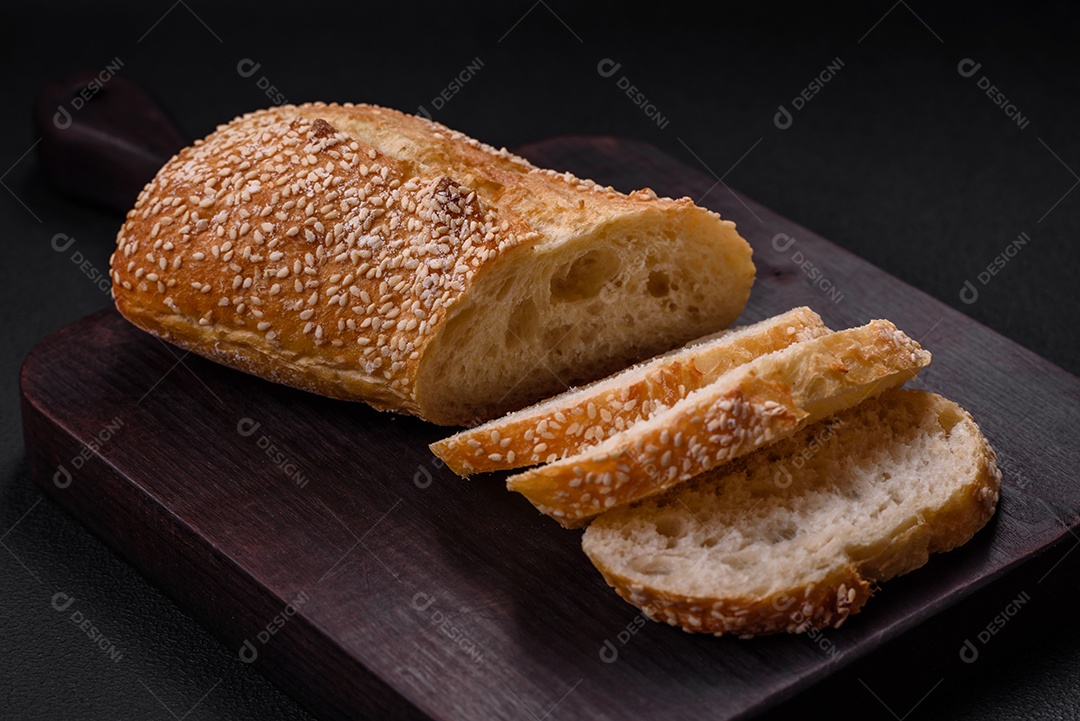 Baguete de farinha de trigo crocante com sementes de gergelim em uma tábua de madeira sobre um fundo escuro de concreto