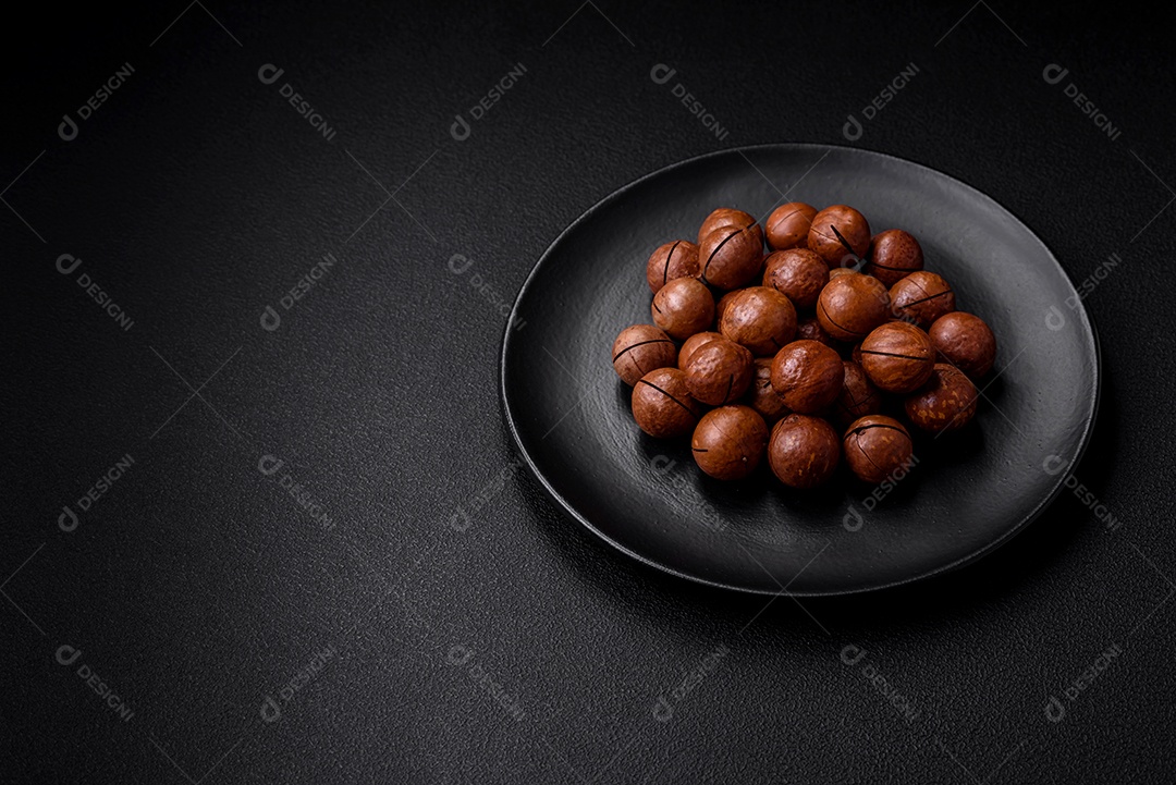 Deliciosas nozes de macadâmia torradas com casca sobre um fundo de concreto texturizado escuro. Comida vegetariana