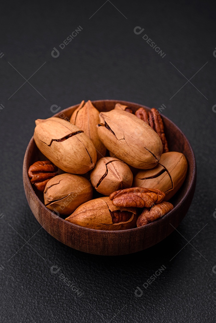 Nozes pecan com casca e descascadas em uma tigela redonda de madeira sobre um fundo escuro de concreto