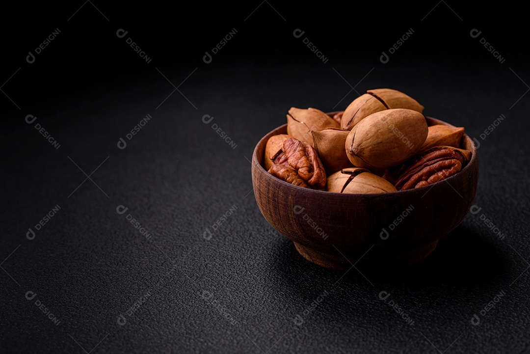 Nozes pecan com casca e descascadas em uma tigela redonda de madeira sobre um fundo escuro de concreto