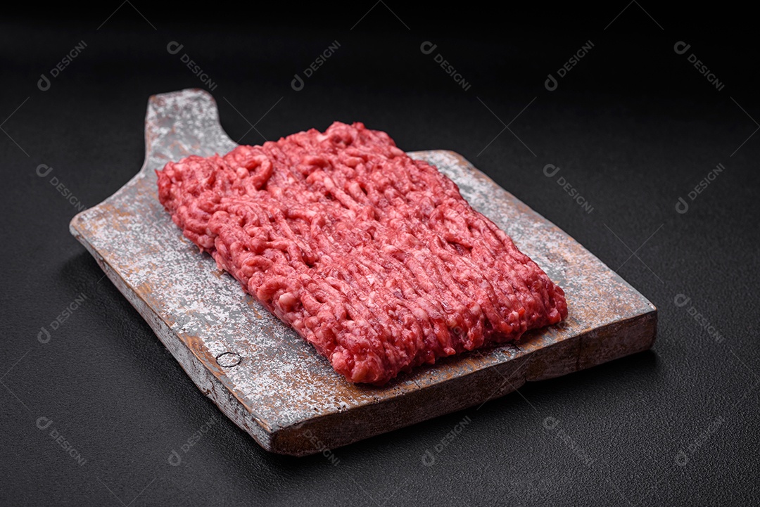 Carne moída crua ou carne de porco em uma tábua de madeira com especiarias e sal sobre um fundo escuro de concreto