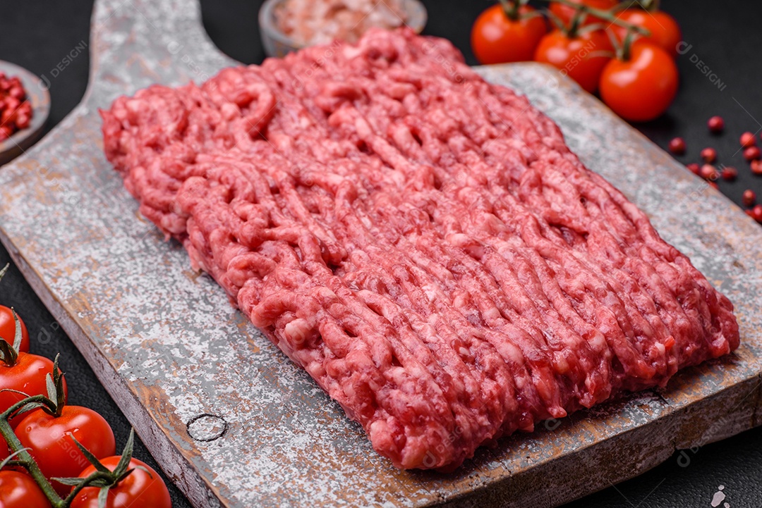 Carne moída crua ou carne de porco em uma tábua de madeira com especiarias e sal sobre um fundo escuro de concreto
