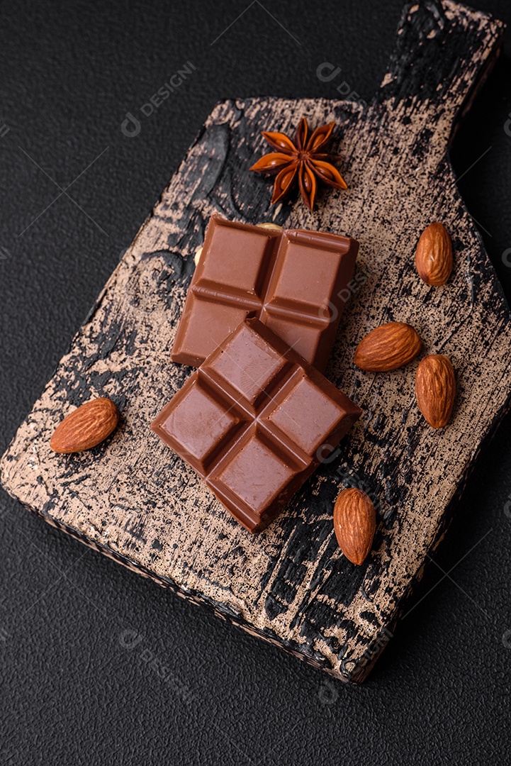 Delicioso chocolate preto doce quebrado em cubos em uma tábua de madeira sobre um fundo escuro de concreto