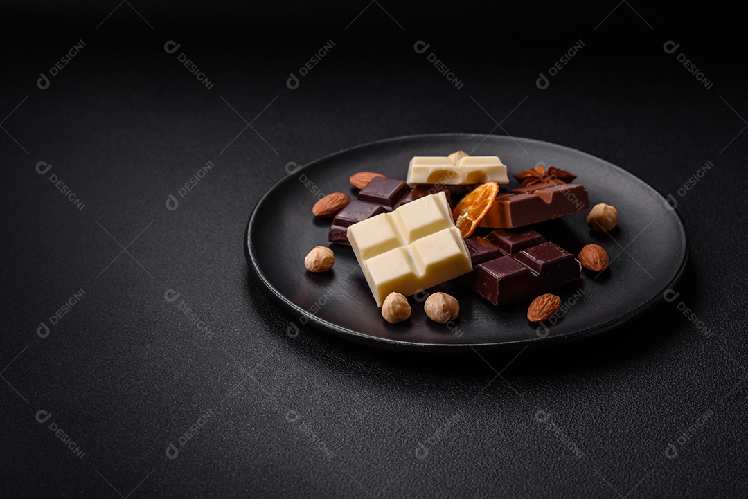 Uma mistura de vários tipos de delicioso chocolate doce quebrado em cubos em uma placa preta sobre fundo escuro de concreto