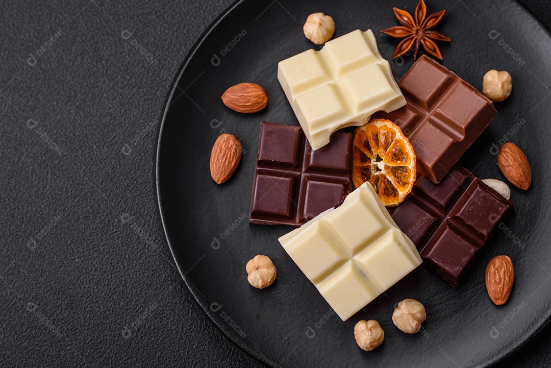 Uma mistura de vários tipos de delicioso chocolate doce quebrado em cubos em uma placa preta sobre fundo escuro de concreto