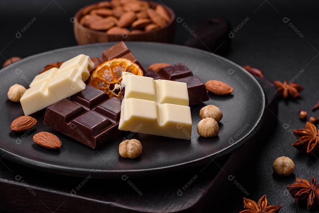 Uma mistura de vários tipos de delicioso chocolate doce quebrado em cubos em uma placa preta sobre fundo escuro de concreto