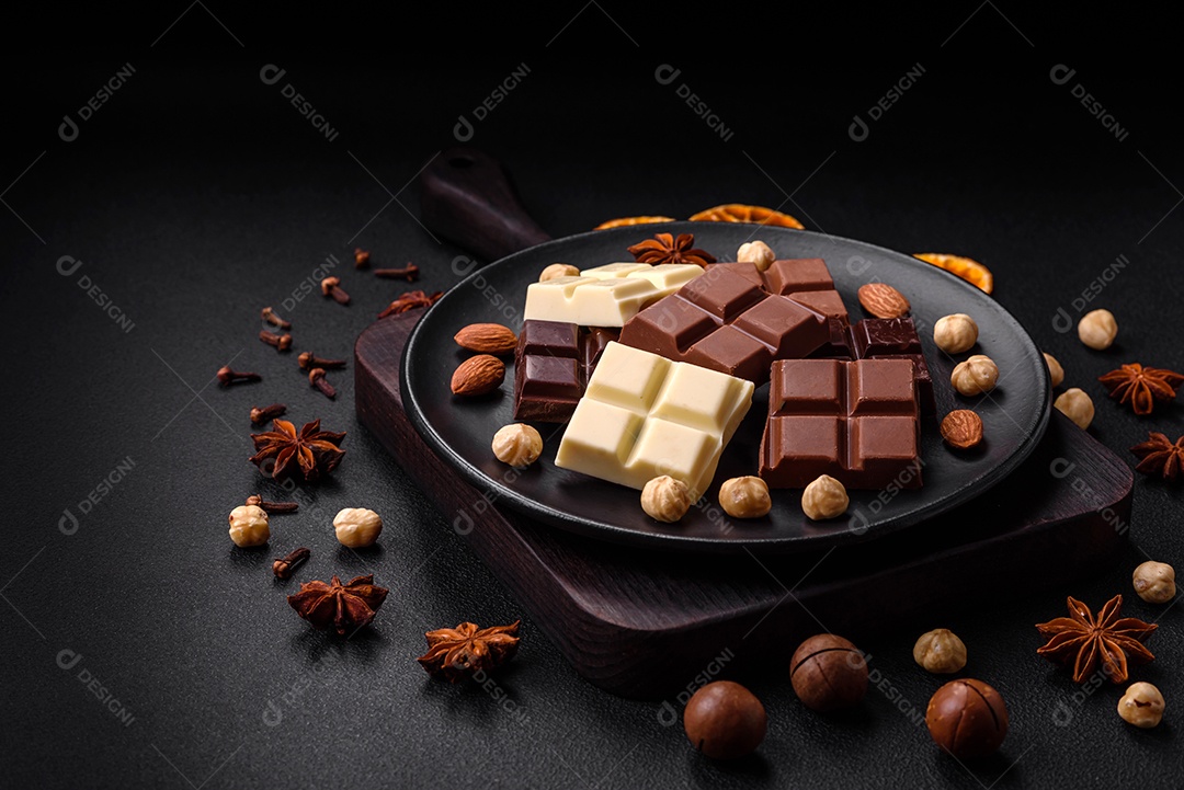 Uma mistura de vários tipos de delicioso chocolate doce quebrado em cubos em uma placa preta sobre fundo escuro de concreto