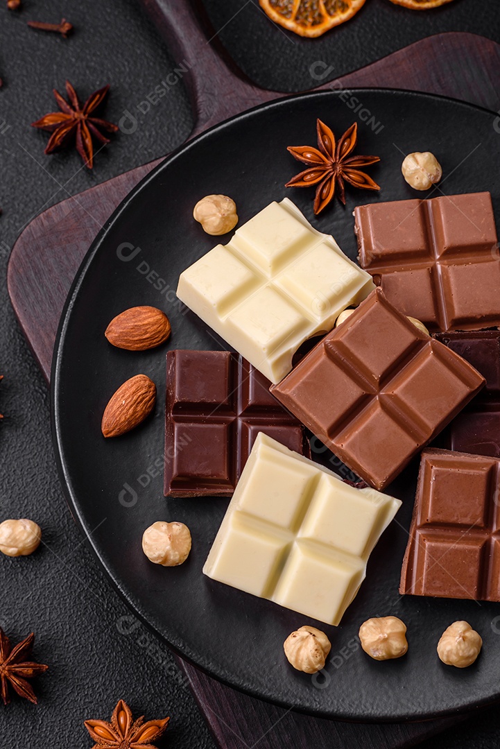 Uma mistura de vários tipos de delicioso chocolate doce quebrado em cubos em uma placa preta sobre fundo escuro de concreto