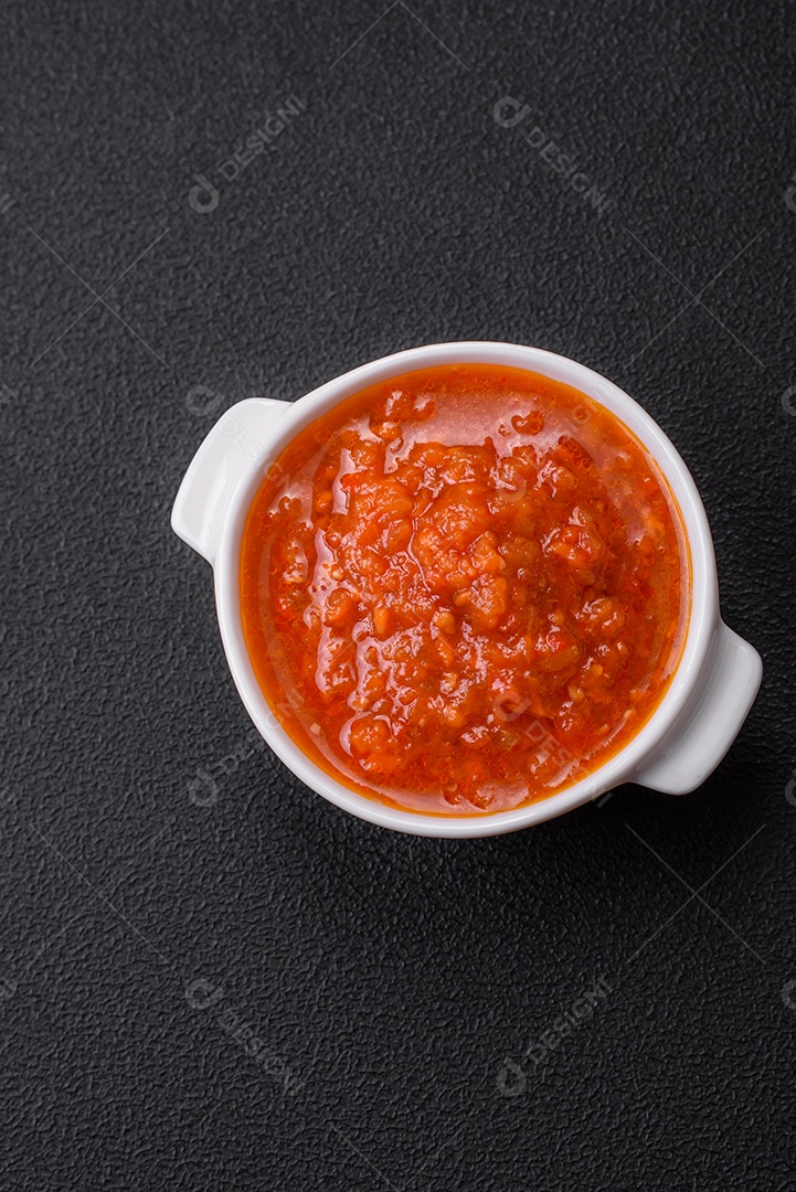 Delicioso molho de tomate picante com pimenta, alho, sal, especiarias e ervas em uma molheira branca sobre um fundo escuro de concreto