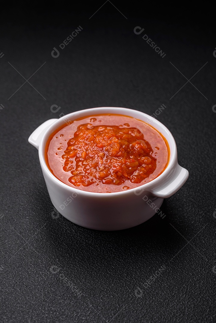 Delicioso molho de tomate picante com pimenta, alho, sal, especiarias e ervas em uma molheira branca sobre um fundo escuro de concreto
