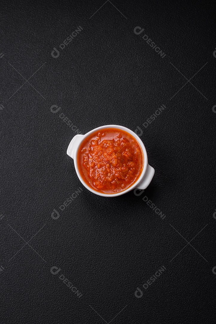 Delicioso molho de tomate picante com pimenta, alho, sal, especiarias e ervas em uma molheira branca sobre um fundo escuro de concreto