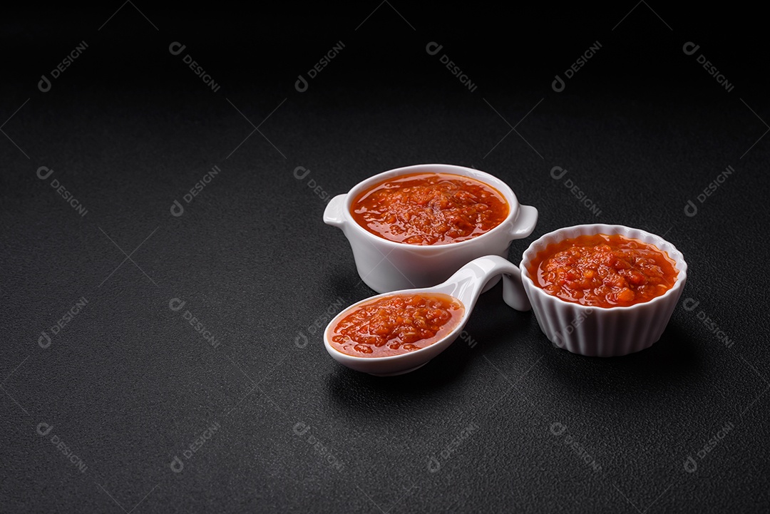 Delicioso molho de tomate picante com pimenta, alho, sal, especiarias e ervas em uma molheira branca sobre um fundo escuro de concreto