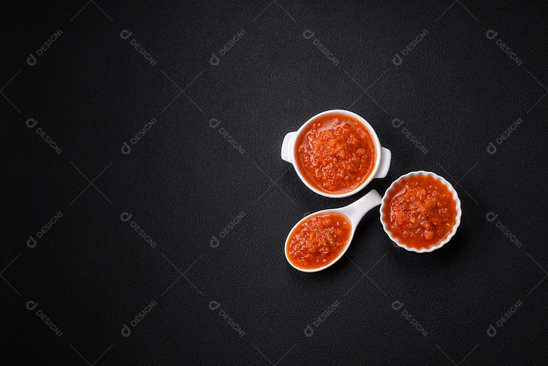 Delicioso molho de tomate picante com pimenta, alho, sal, especiarias e ervas em uma molheira branca sobre um fundo escuro de concreto