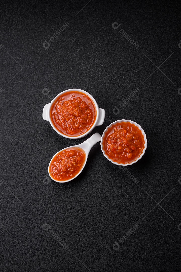 Delicioso molho de tomate picante com pimenta, alho, sal, especiarias e ervas em uma molheira branca sobre um fundo escuro de concreto