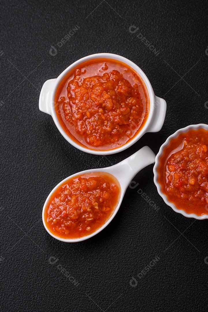 Delicioso molho de tomate picante com pimenta, alho, sal, especiarias e ervas em uma molheira branca sobre um fundo escuro de concreto