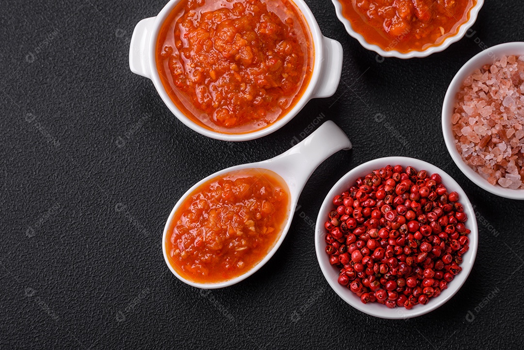 Delicioso molho de tomate picante com pimenta, alho, sal, especiarias e ervas em uma molheira branca sobre um fundo escuro de concreto