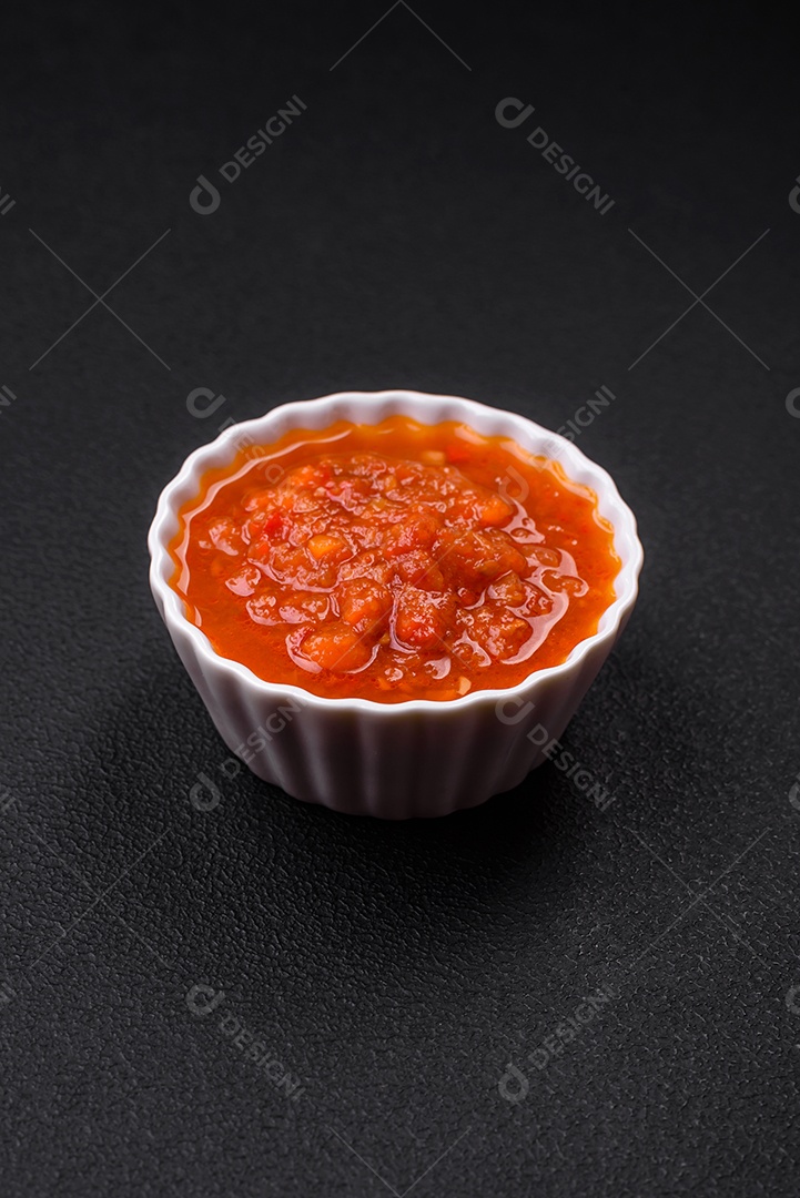 Delicioso molho de tomate picante com pimenta, alho, sal, especiarias e ervas em uma molheira branca sobre um fundo escuro de concreto