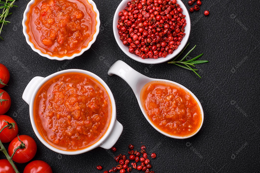 Delicioso molho de tomate picante com pimenta, alho, sal, especiarias e ervas em uma molheira branca sobre um fundo escuro de concreto