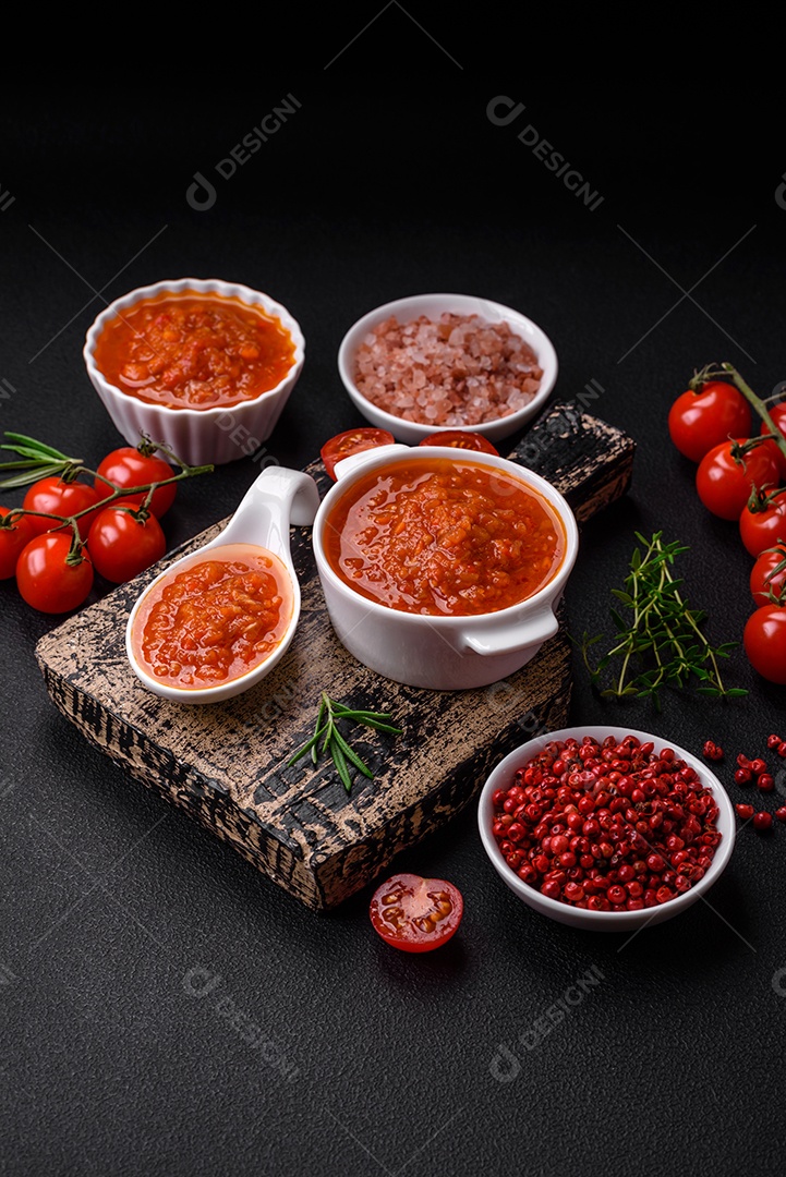 Delicioso molho de tomate picante com pimenta, alho, sal, especiarias e ervas em uma molheira branca sobre um fundo escuro de concreto