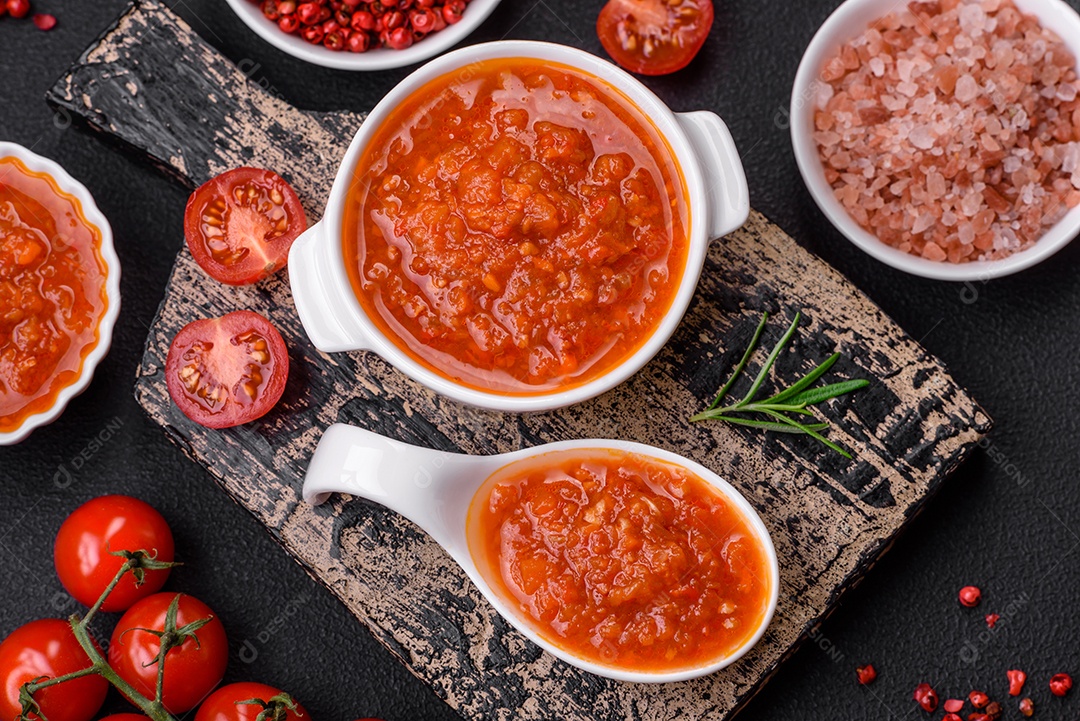Delicioso molho de tomate picante com pimenta, alho, sal, especiarias e ervas em uma molheira branca sobre um fundo escuro de concreto